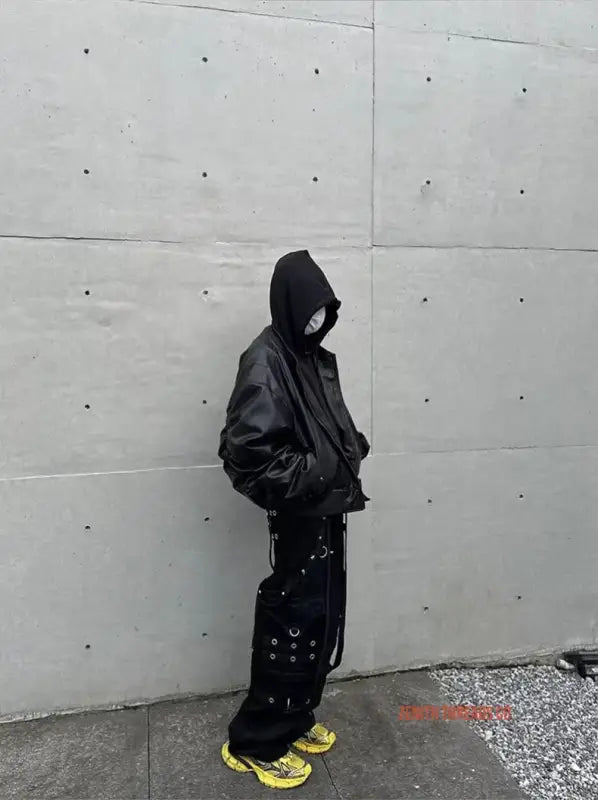 Hooded figure in dark clothing and bright yellow sneakers leaning against a wall.