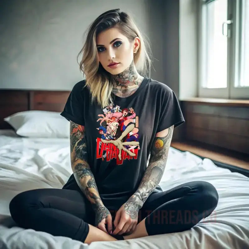 A person with tattoos wearing a black graphic t-shirt and dark pants sitting cross-legged on a bed.