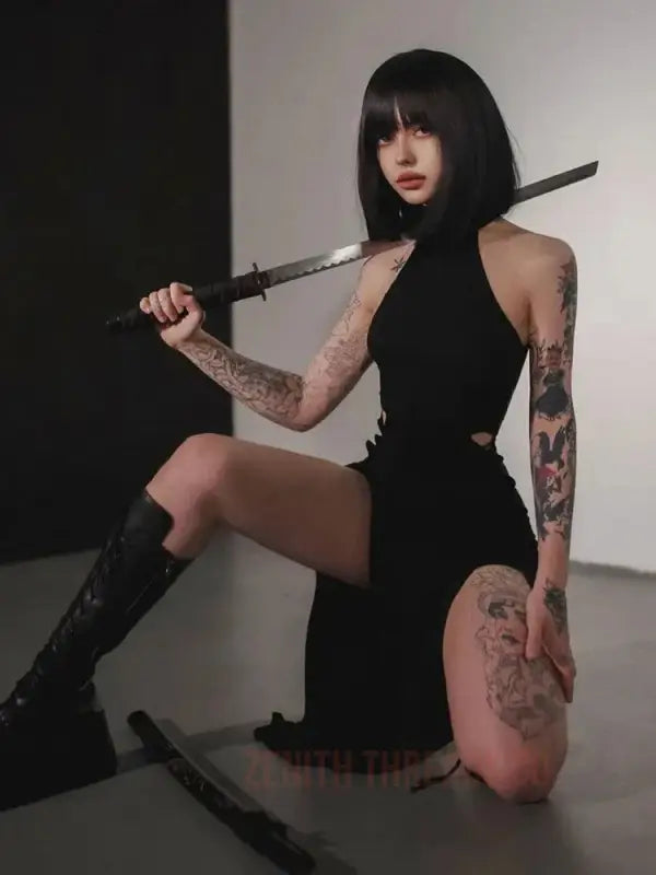 A tattooed person in a black halter dress and boots posing with a sword.