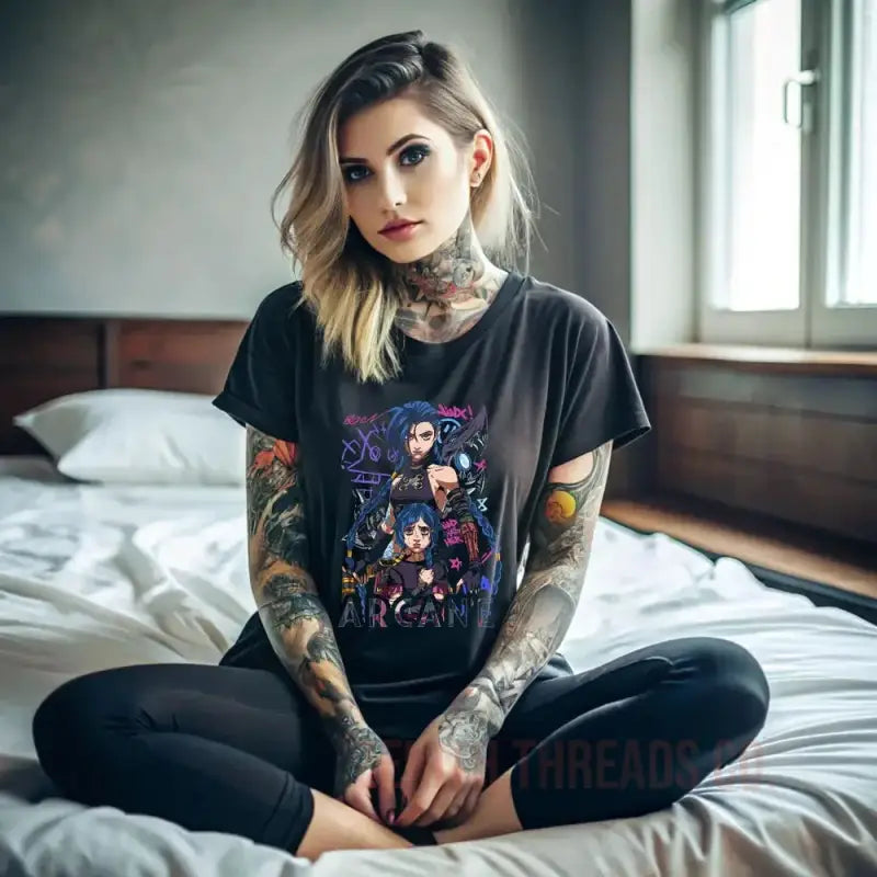 Person wearing a black graphic t-shirt with colorful tattoos sitting cross-legged on a bed.