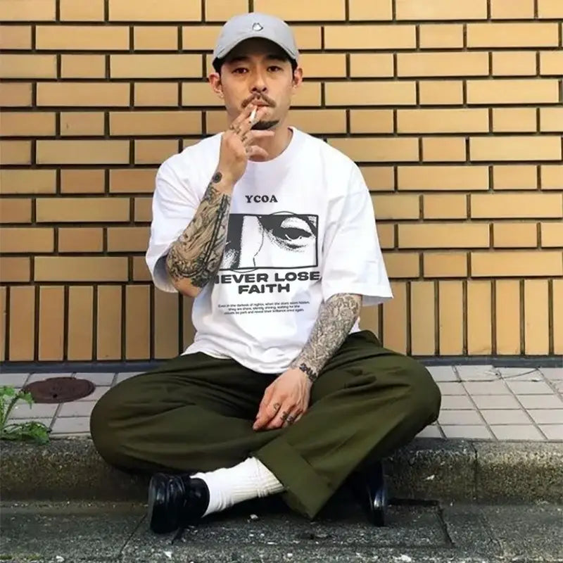 Someone wearing a white graphic t-shirt, olive green pants, and a gray baseball cap sits cross-legged while smoking.