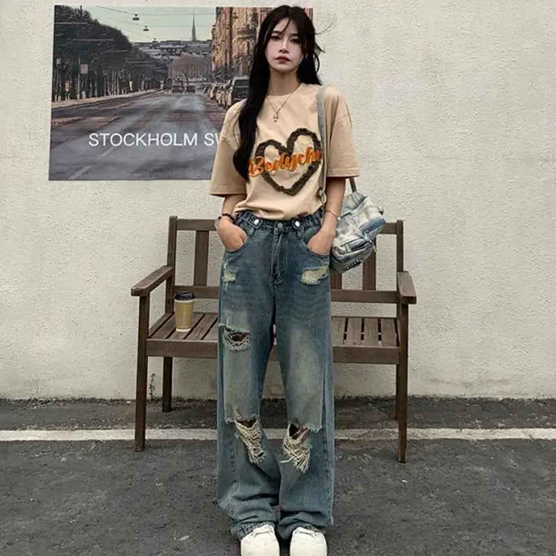 A person wearing a beige graphic t-shirt, distressed baggy jeans, and white sneakers.