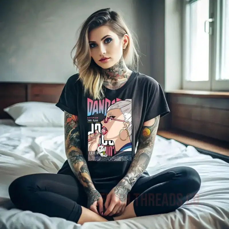 Person wearing a black graphic t-shirt and tattoos sitting cross-legged on a bed.